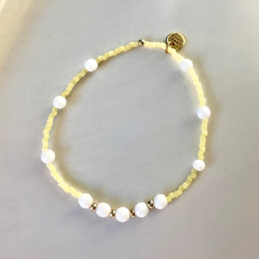 A delicate beaded bracelet with pale yellow micro-beads, white round beads, and small gold spacer beads, displayed on a light gray surface.