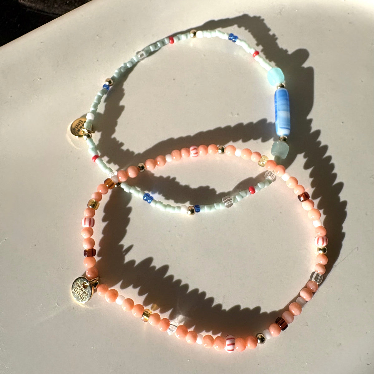 Two beaded stretch bracelets with gold-toned charms, displayed on a white surface in bright natural sunlight. One bracelet is light blue with a blue cylindrical bead, and the other is peach-pink with a round gold charm.