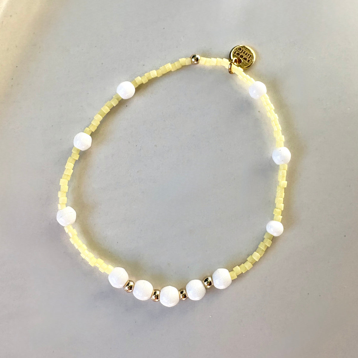 A delicate beaded bracelet with pale yellow micro-beads, white round beads, and small gold spacer beads, displayed on a light gray surface.