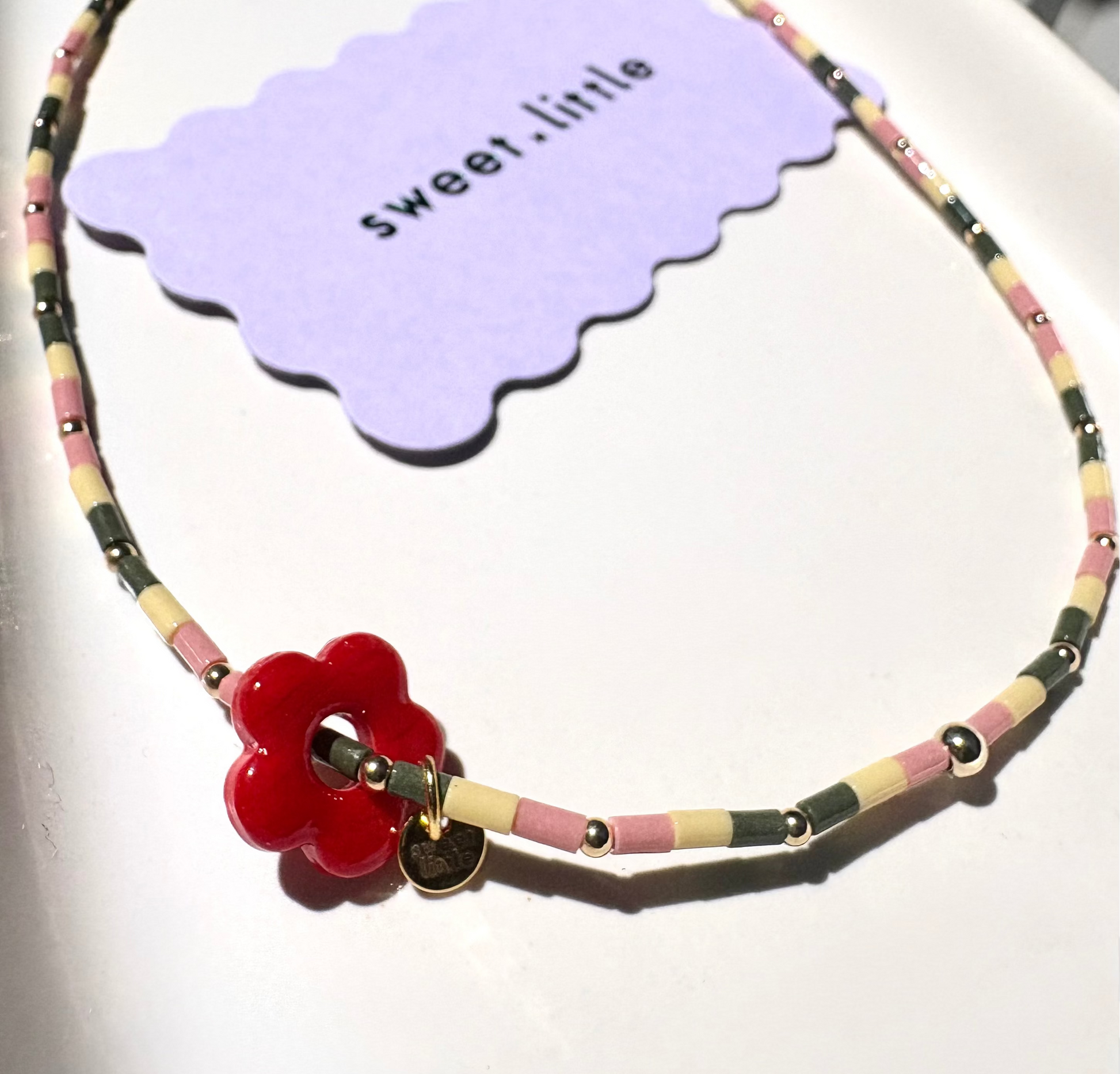 Necklace with pink, yellow and cream beads with a red flower pendant on a white surface, featuring a 'sweet little' tag.