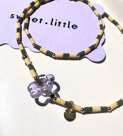 Necklace with green and cream beads and a purple flower pendant on a white tray with 'sweet little' text.